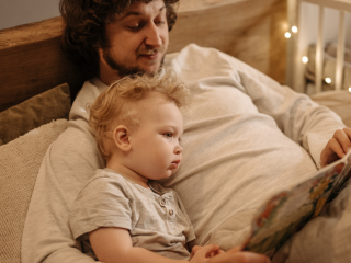 Dad, lying back on his bed, with his young blond son snuggled in to him. Together they are looking at a book.