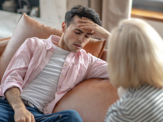 Young man, sitting on a sofa, with his eyes closed and his forehead resting on his hand. He is wearing a pink shirt, a white T-shirt and blue jeans. Sitting opposite him, with his back to the camera, is a blonde-haired woman. It looks as if he is in distress and talking to her.