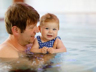 Dad holding his baby daughter in the water at their local swimming pool. Baby girl is confidently looking at the camera, dad is watching her face and smiling.